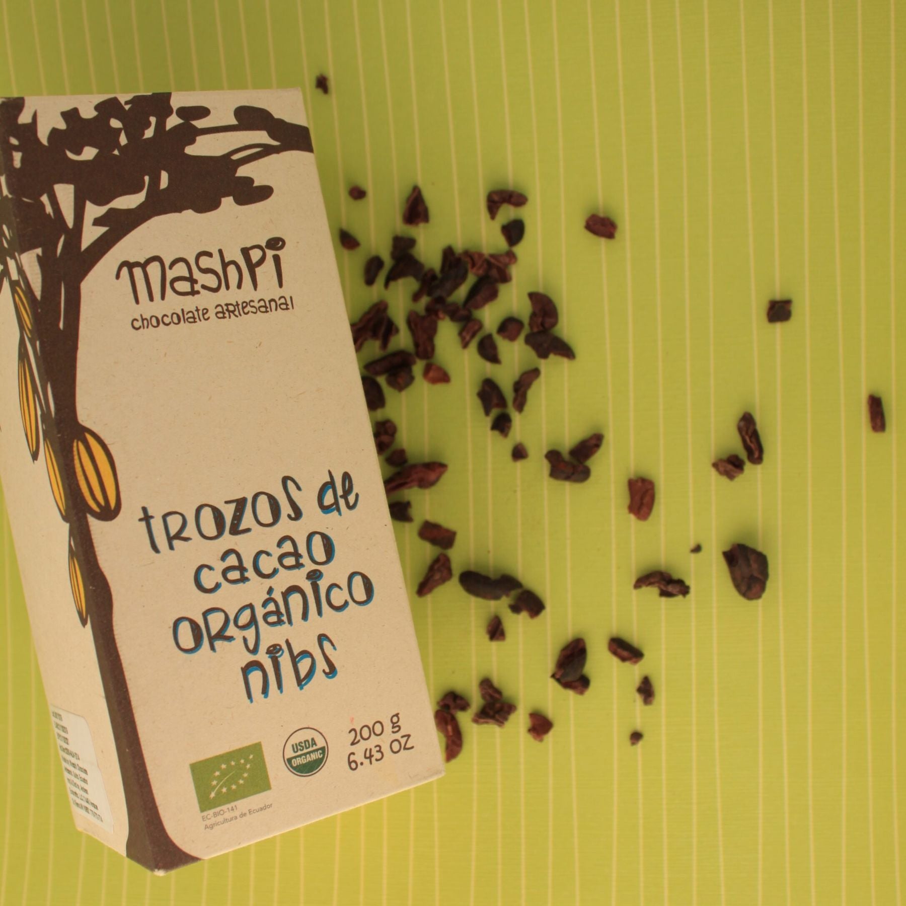 Box of Mashpi Chocolate Artesenal Organic Cacao Nibs with cacao nibs sprinkled across a green striped background.